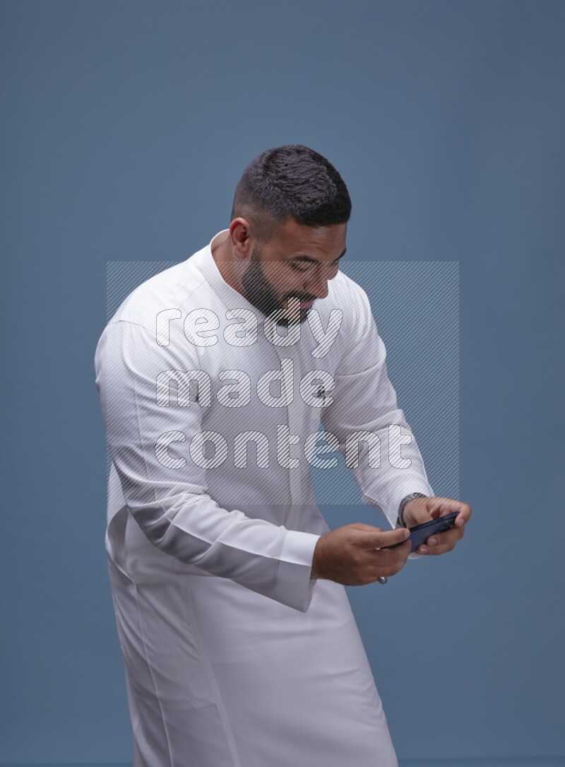A man Playing a Game on his Smartphone  on Blue Background wearing Saudi Thob