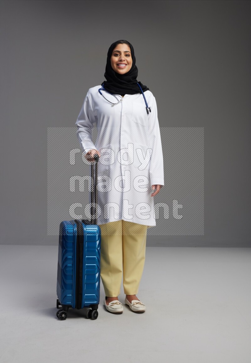Saudi woman wearing lab coat with stethoscope standing holding Travel bag on Gray background