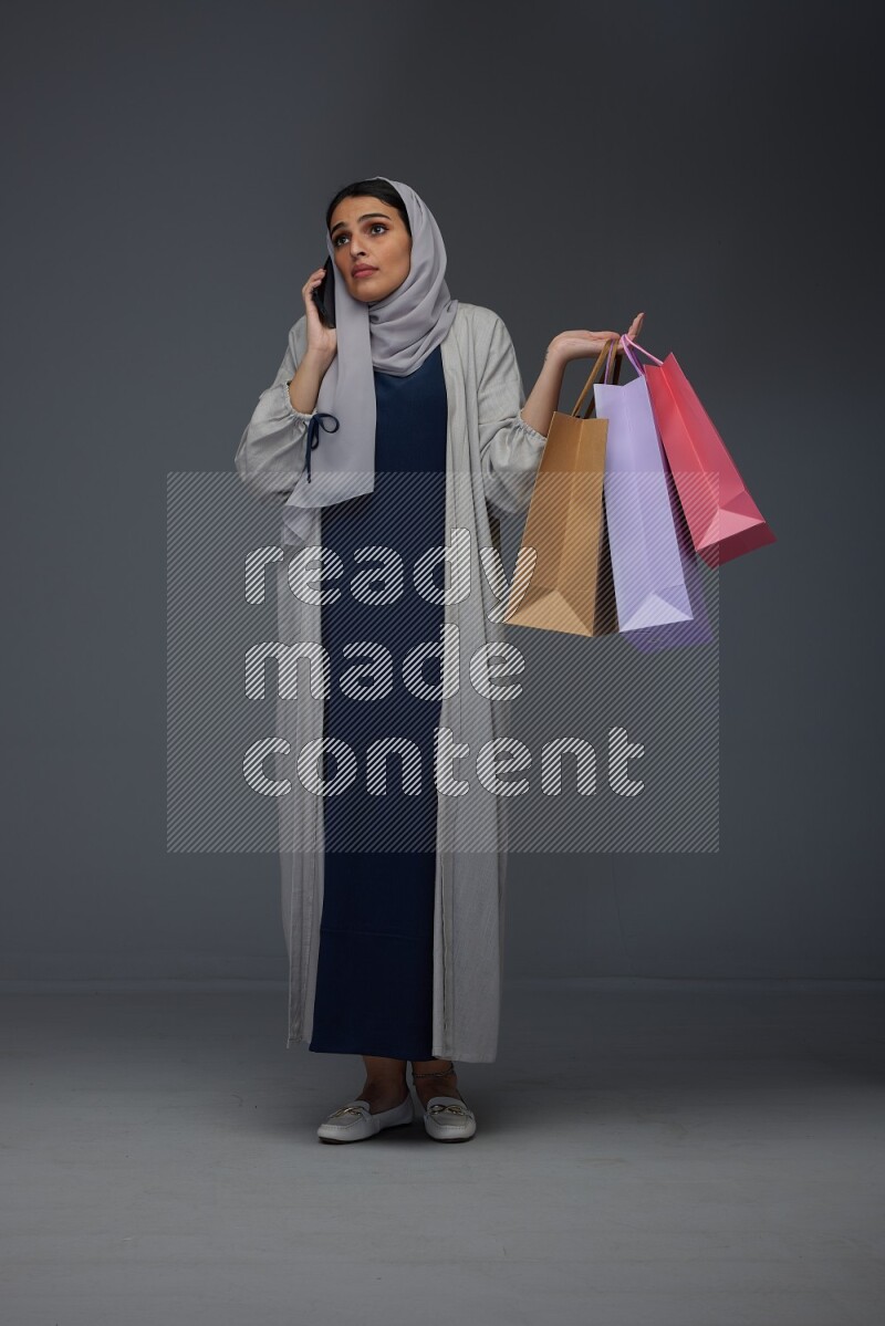 A Saudi woman wearing a light gray Abaya and head scarf standing and holding shopping bags making different poses eye level on a grey background