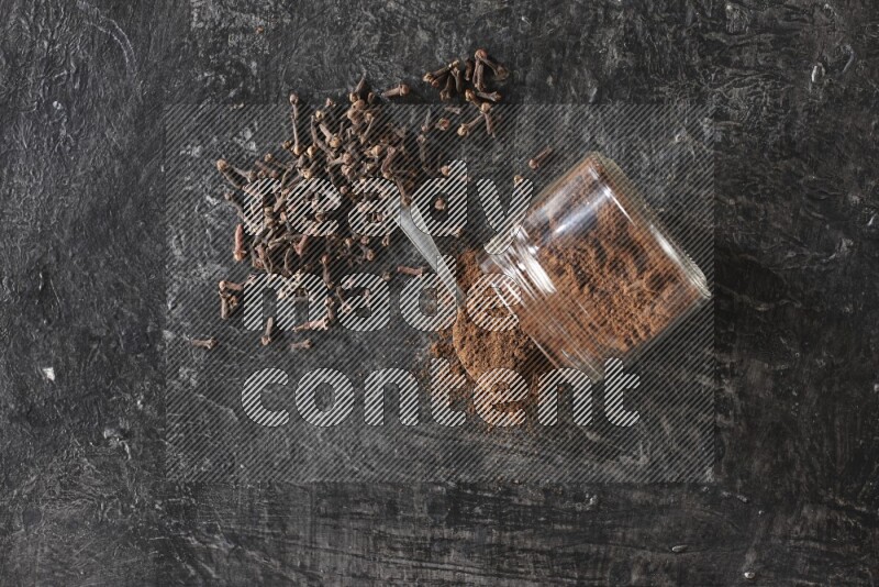 A flipped glass jar and metal spoon full of cloves powder with cloves spread on a textured black flooring