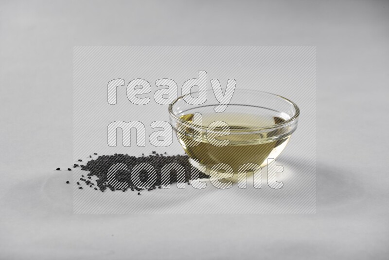 A glass bowl full of black seeds oil with black seeds beside it on a white flooring