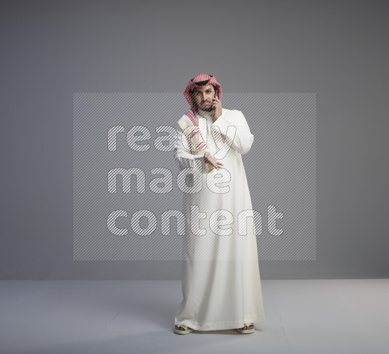 A Saudi man standing wearing thob and red shomag talking on phone on gray background