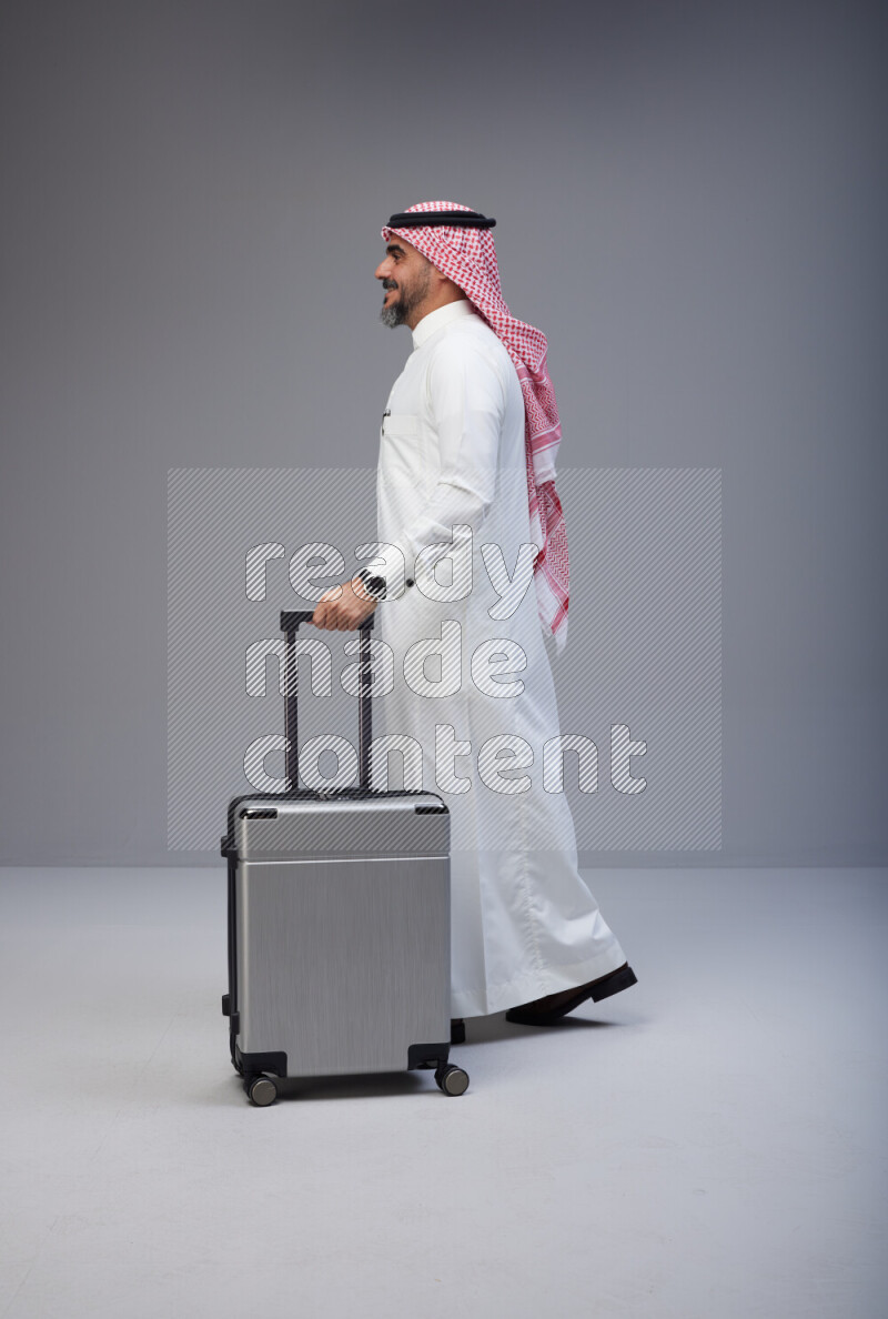 Saudi man wearing Thob and red Shomag standing holding Travel bag on Gray background