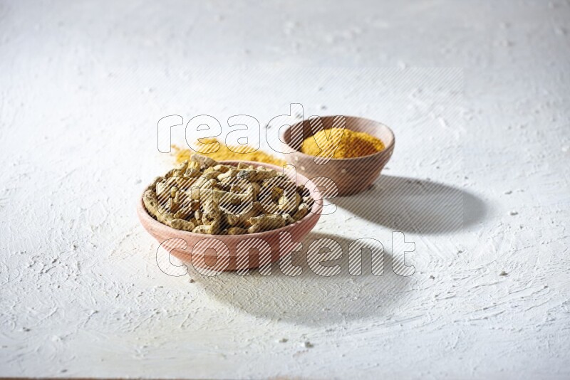 2 wooden bowls, one full of turmeric powder and the other full of dried turmeric whole fingers on a textured white flooring