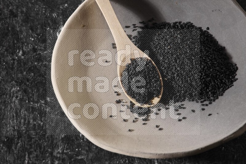 A multicolored pottery plate full of black seeds and wooden spoon full of seeds on a textured black flooring