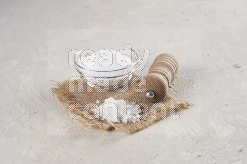 A glass bowl full of white salt with a wooden grinder on a burlap fabric all on white background