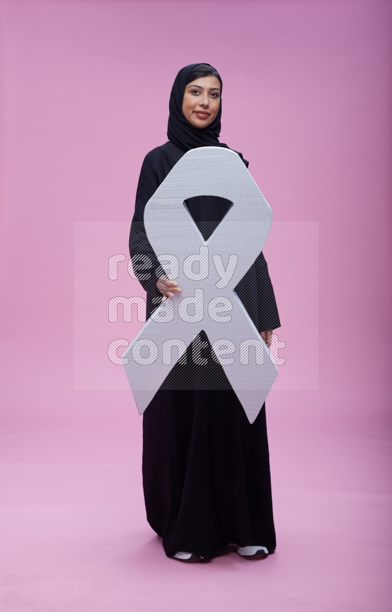 Saudi woman wearing pink ribbon on abaya standing holding awareness ribbon on plain pink background