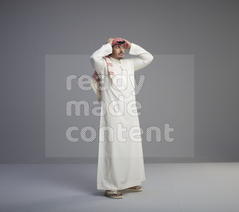 A Saudi man standing wearing thob and red shomag interacting with the camera on gray background