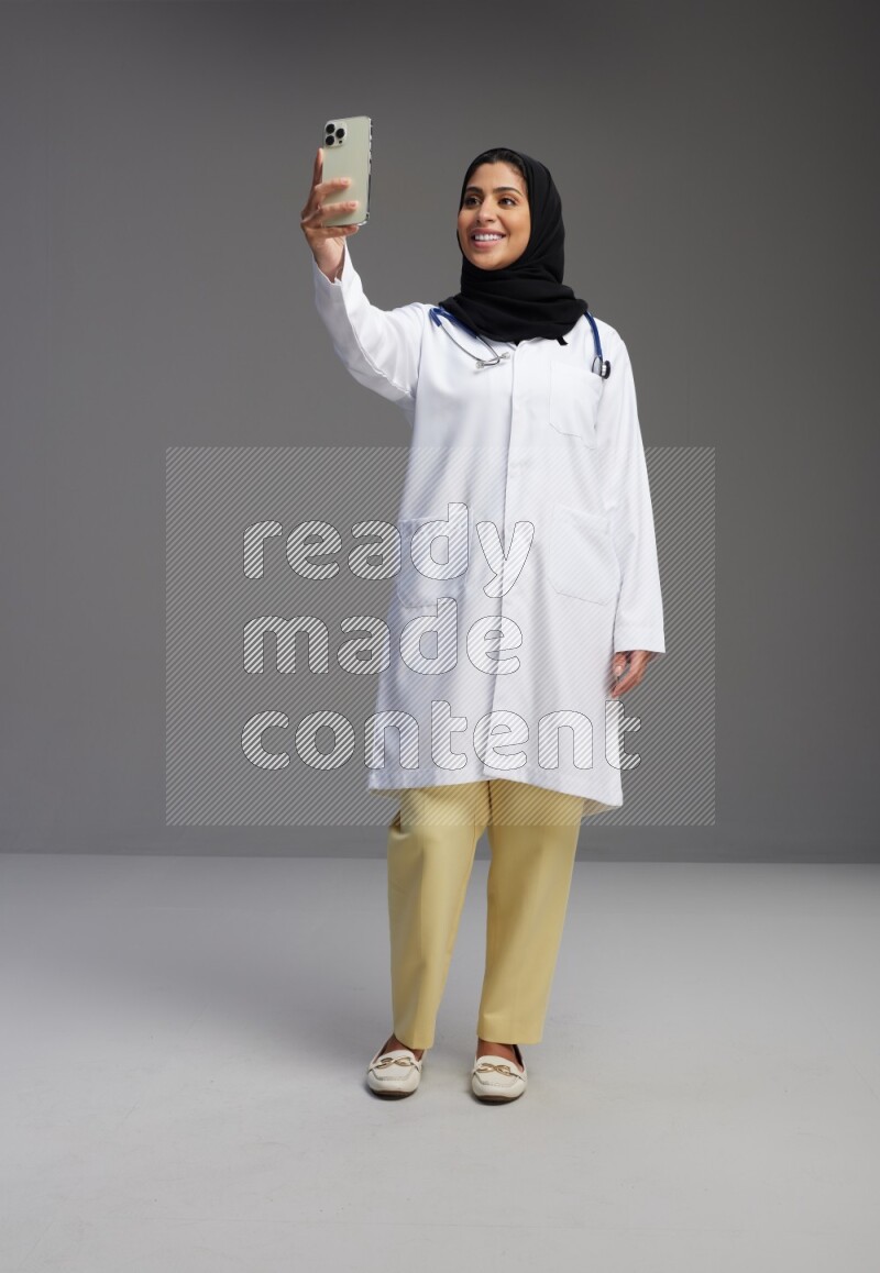 Saudi woman wearing lab coat with stethoscope standing taking selfie on Gray background