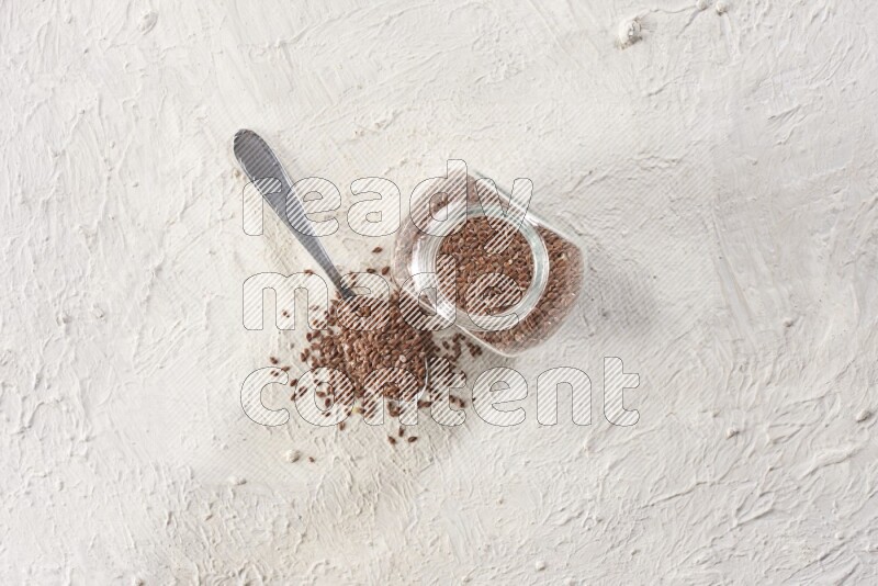A glass spice jar full of flax seeds and a metal spoon full of the seeds on a textured white flooring