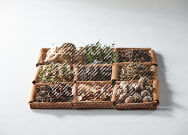 9 squares of cinnamon sticks full of tea in the middle surrounded by nutmeg, dried mint, cloves, dried basil, dried ginger, cinnamon, star anise and cardamom on white flooring