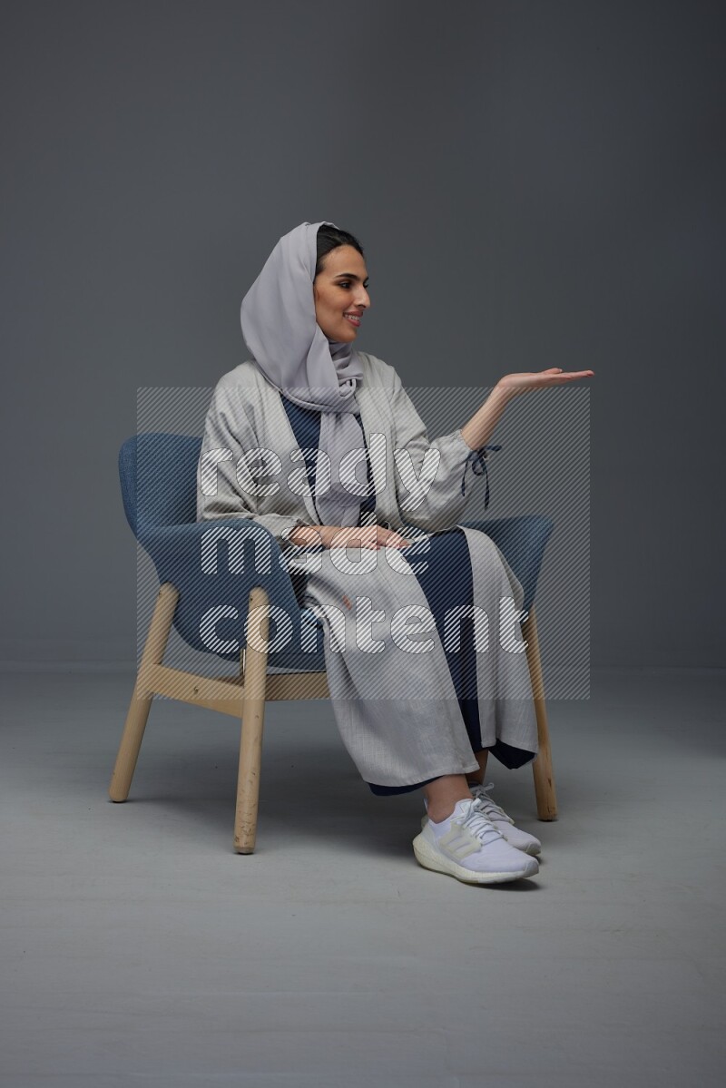 A Saudi woman wearing a light gray Abaya and head scarf sitting on a dark grey chair while making multi hand gestures eye level on a grey background