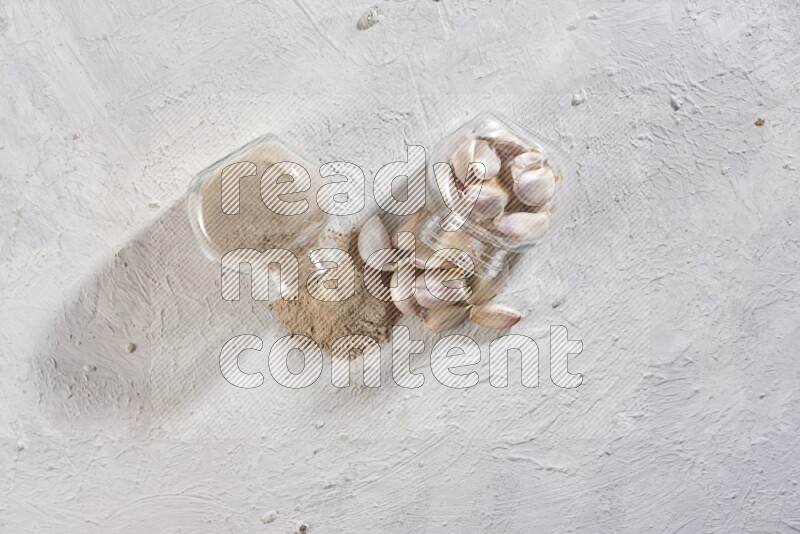 وعاءان للبهارات من الزجاج احدهما ممتلئ ببودرة الثوم والأخر ممتلئ بفصوص الثوم وكلاهما مقلوب علي خلفية بيضاء