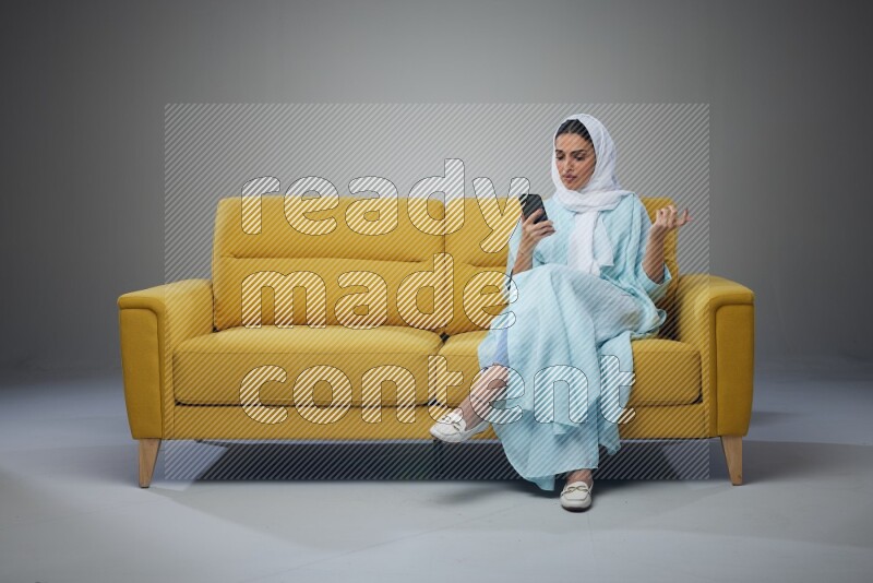 A Saudi woman wearing a light blue Abaya and white head scarf sitting on a yellow sofa and using her phone eye level on a grey background