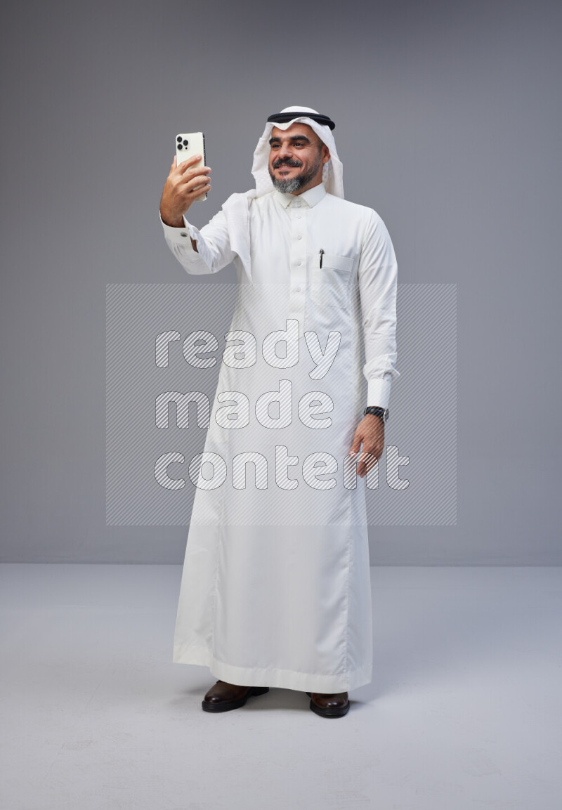Saudi man Wearing Thob and white Shomag standing taking selfie on Gray background