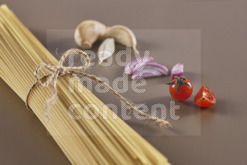 Raw pasta with different ingredients such as cherry tomatoes, garlic, onions, red chilis, black pepper, white pepper, bay laurel leaves, rosemary and cardamom on beige background