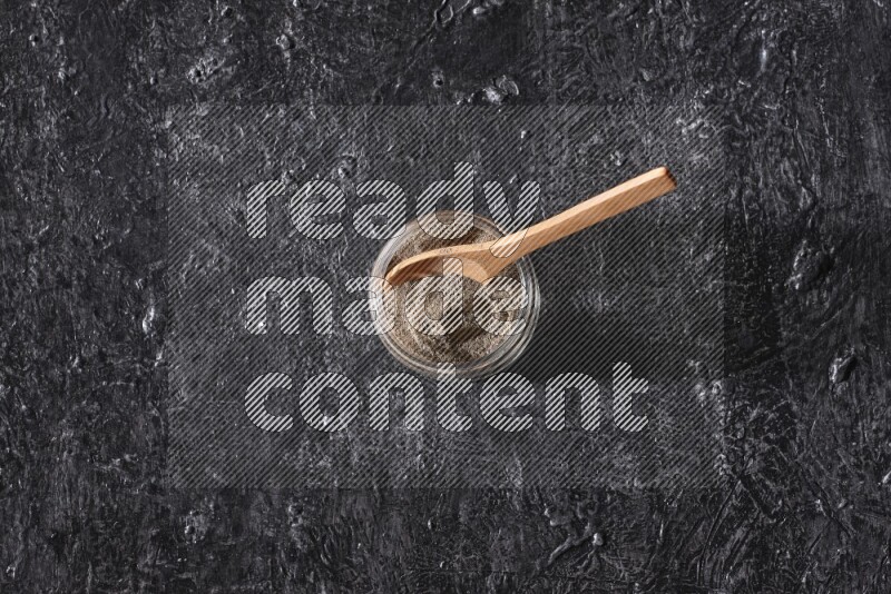 A glass jar full of black pepper powder and a wooden spoon on a textured black flooring