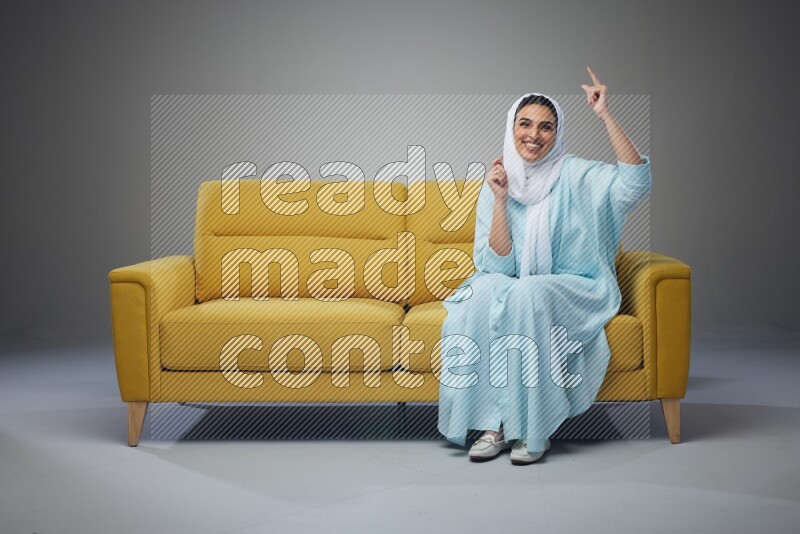 A Saudi woman wearing a light blue Abaya and white head scarf sitting on a yellow sofa and thinking and pointing up eye level on a grey background