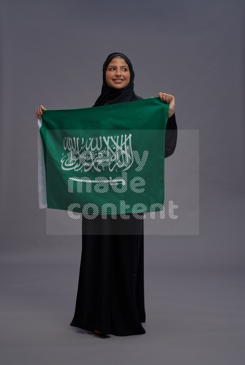 Saudi woman wearing Abaya standing holding Saudi flag on gray background
