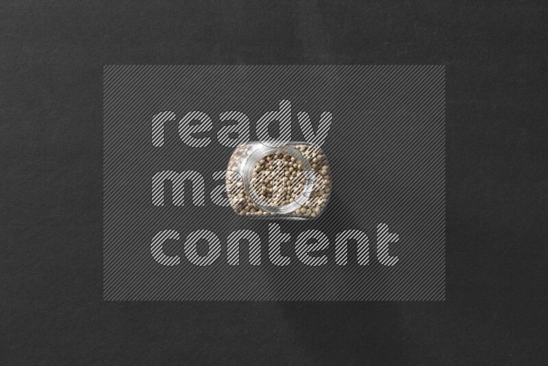 A glass spice jar full of white peppers on black flooring