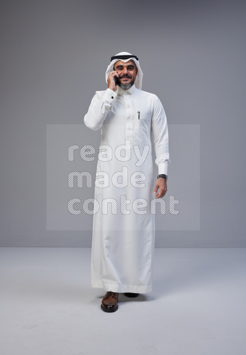 Saudi man Wearing Thob and white Shomag standing talking on phone on Gray background