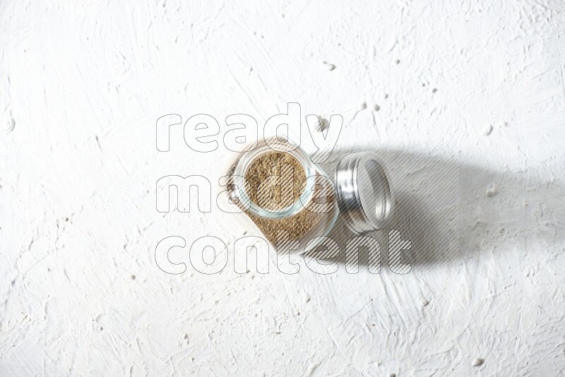 A glass spice jar full of cumin powder on textured white flooring