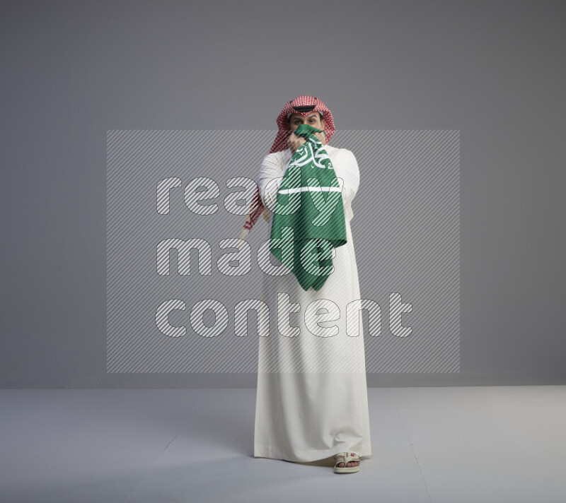 A Saudi man standing wearing thob and red shomag kissing big Saudi flag on gray background
