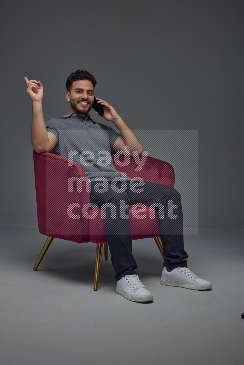 A man wearing casual talking in the phone and making multi hand gestures while sitting on a burgundy chair different angles eye level on a gray background
