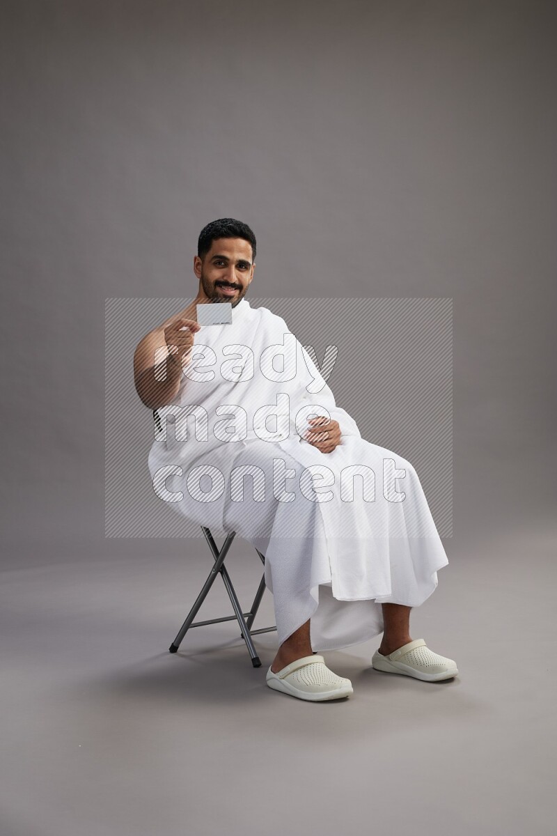 A man wearing Ehram sitting on chair holding ATM card on gray background