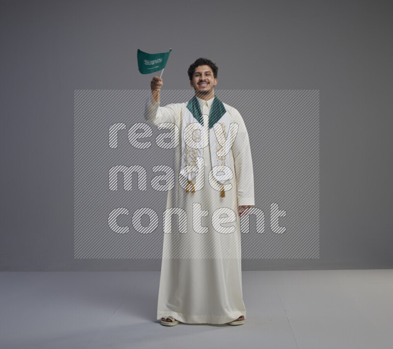 A Saudi man standing wearing thob and saudi flag scarf and holding small saudi flag on gray background