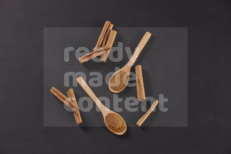 Two wooden spoons full of cinnamon powder with cinnamon sticks on black background