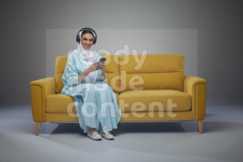 A Saudi woman wearing a light blue Abaya and a white head scarf sitting on a yellow sofa and holding an electronic while wearing headphone eye level on a grey background