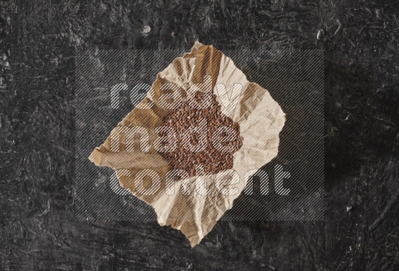 A crumpled piece of paper full of flaxseeds on a textured black flooring