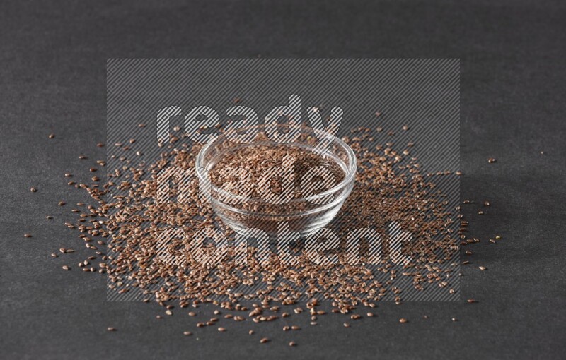A glass bowl full of flaxseeds surrounded by the seeds on a black flooring