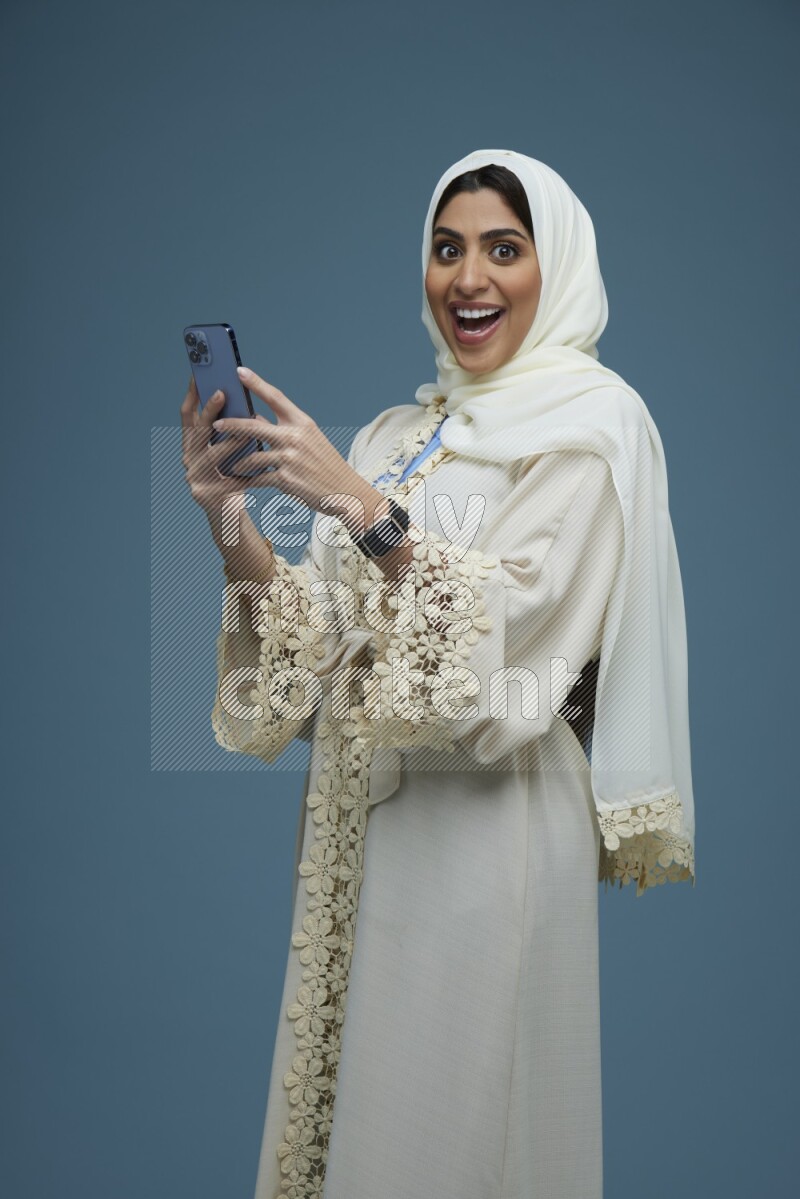 A Saudi woman Texting in a blue background wearing an off-white Abaya Hijab