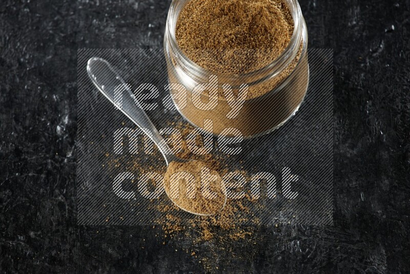 A glass jar and a metal spoon full of cumin powder on a textured black flooring