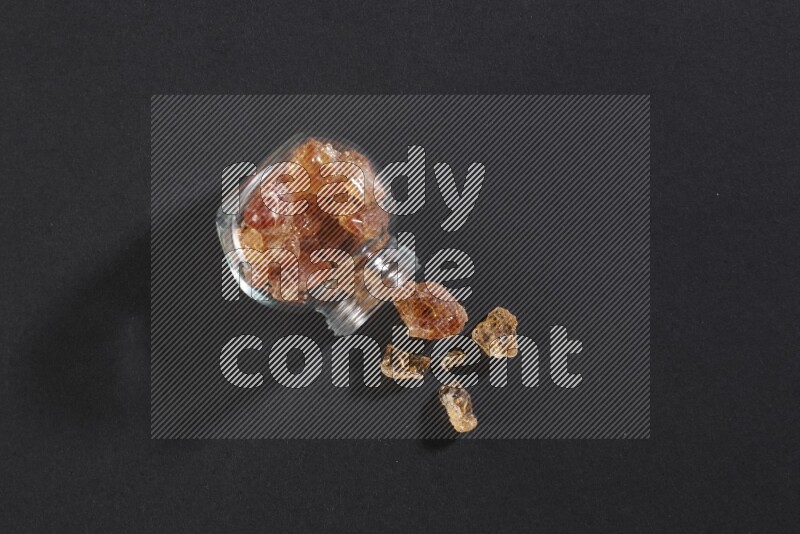 وعاء زجاجي ممتلئ بقطع الصمغ العربي والوعاء مقلوب وبعض القطع متناثرة خارجه علي خلفية سوداء