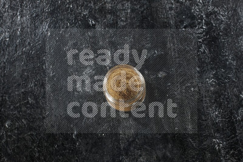 A glass jar full of cumin powder on a textured black flooring