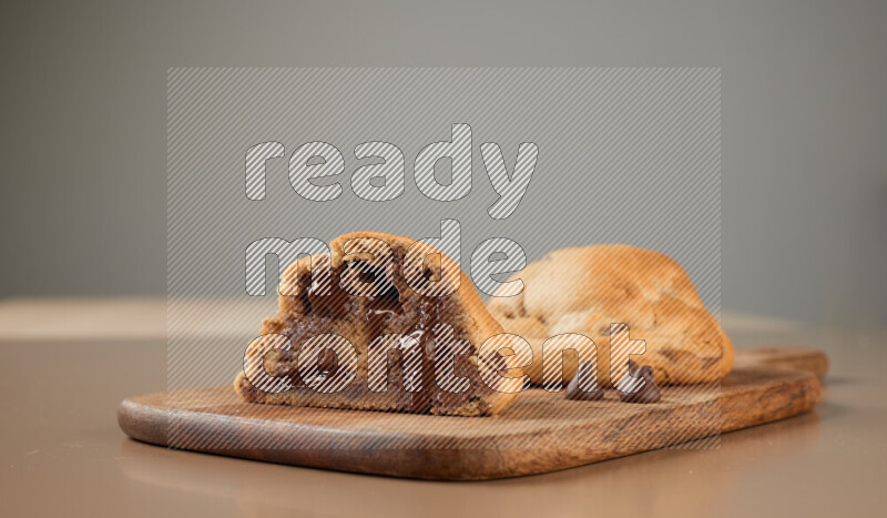 a chocolate chip cookie with another one cut in half on a wooden cutting board on a brown background