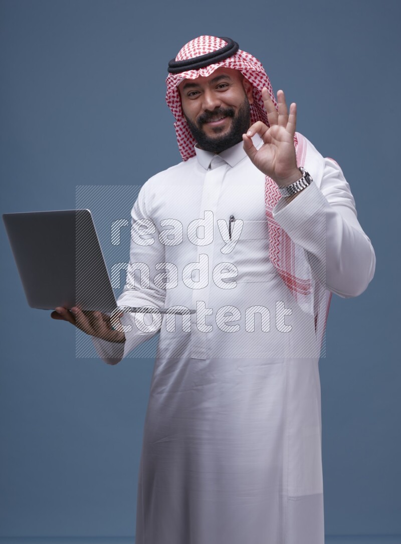 A man standing  with a laptop on Blue Background wearing Saudi Thob and Shomag