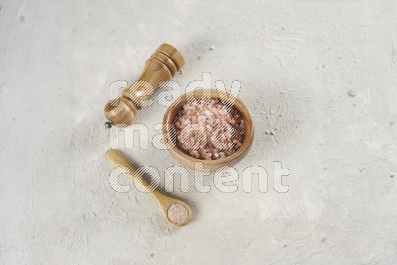 A wooden bowl and spoon filled with coarse pink himalayan salt and a wooden grinder beside them on white background