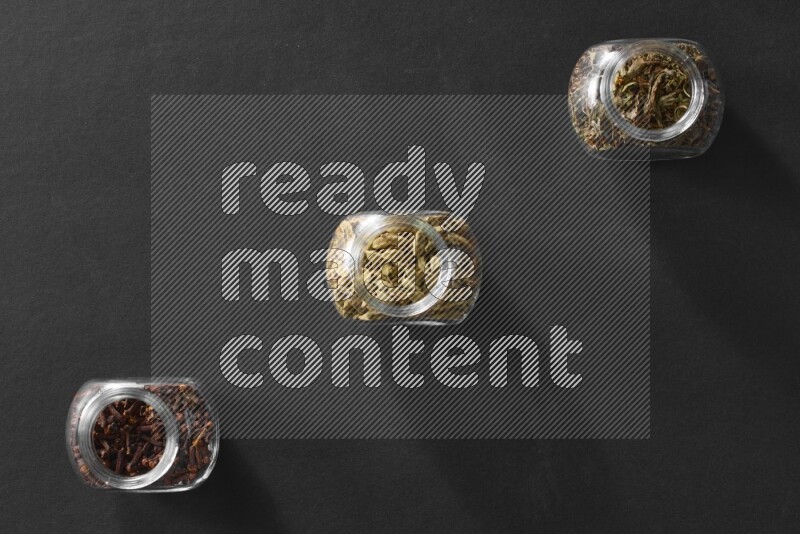 3 glass spice jars full of cloves, cardamom and basil on black flooring