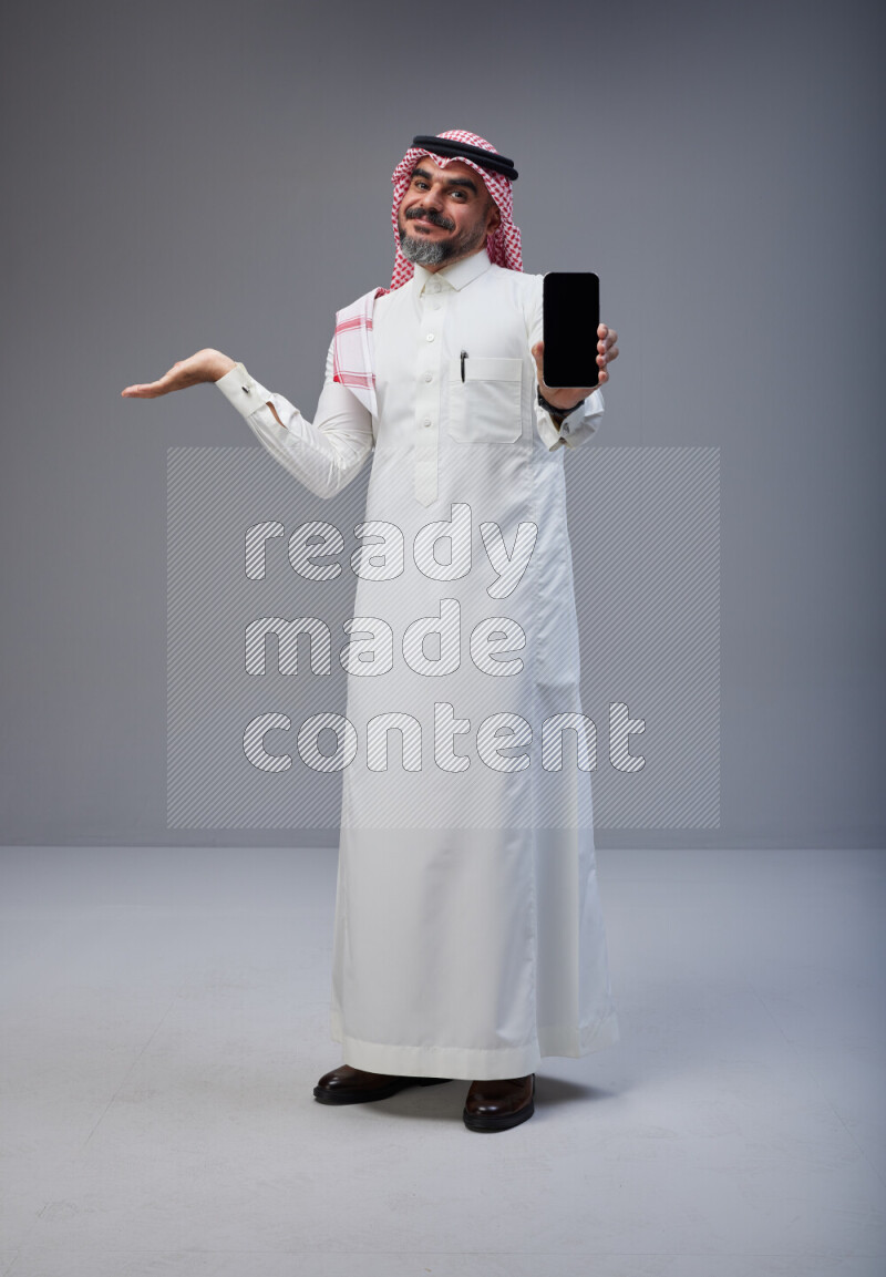 Saudi man Wearing Thob and red Shomag standing showing phone to camera on Gray background