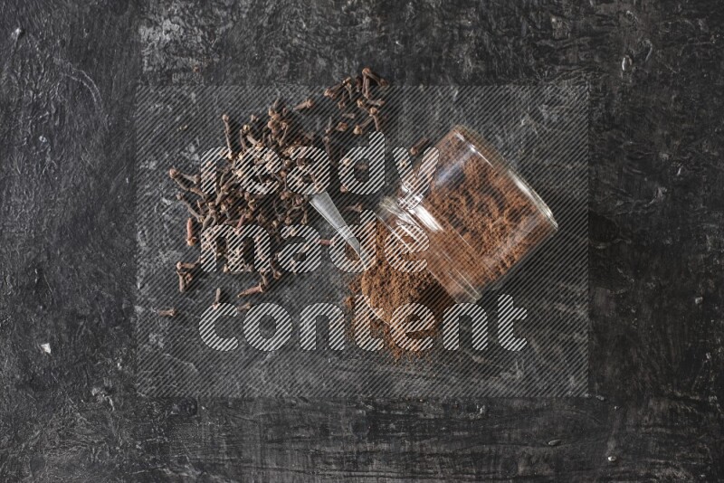 A flipped glass jar and metal spoon full of cloves powder with cloves spread on a textured black flooring
