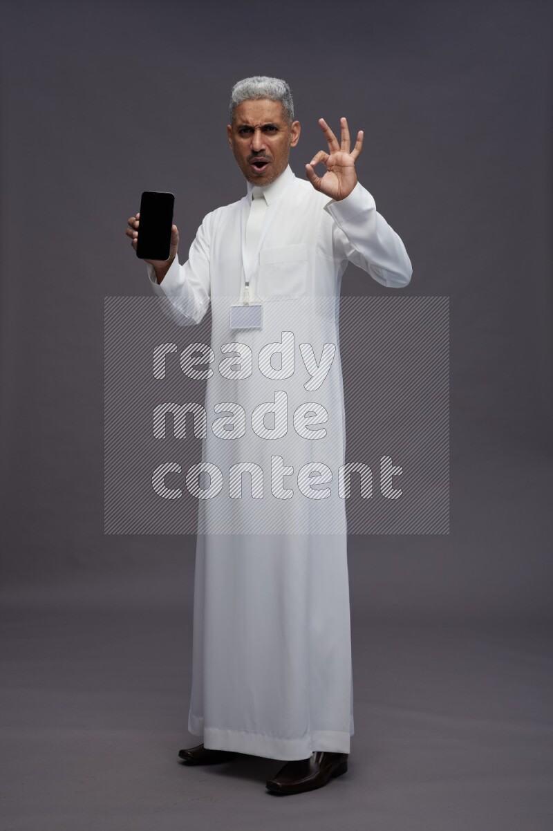 Saudi man wearing thob with neck strap employee badge standing showing phone to camera on gray background
