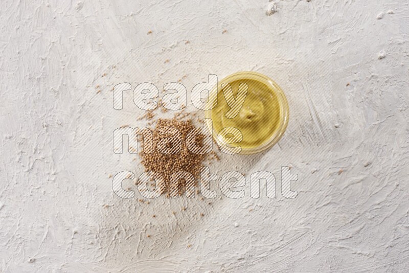 A glass jar full of mustard paste with mustard seeds spread next to it on a textured white flooring