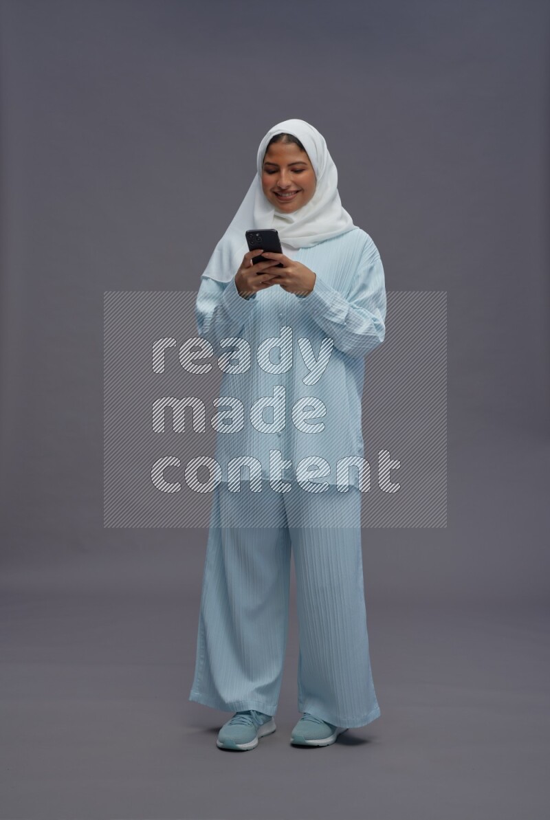 Saudi woman wearing hijab clothes standing texting on phone on gray background