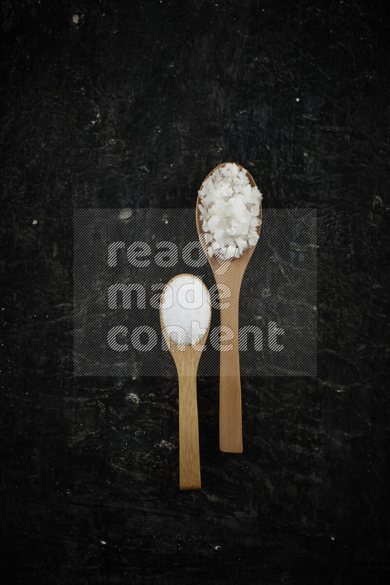 2 wooden spoons filled with fine and coarse salt on black background