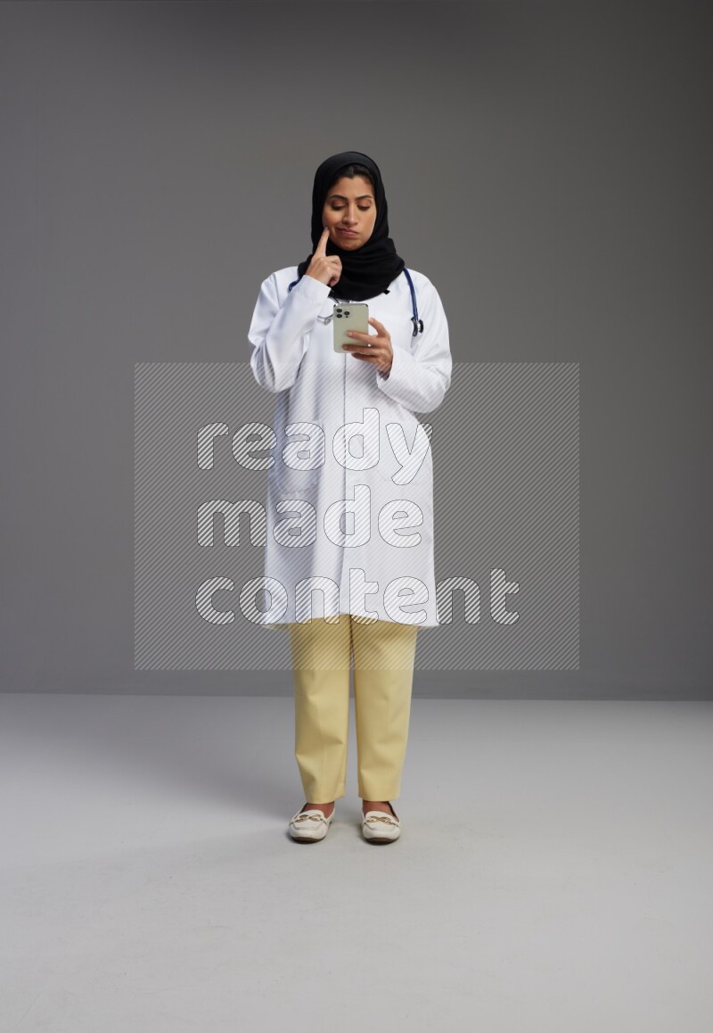 Saudi woman wearing lab coat with stethoscope standing texting on phone on Gray background