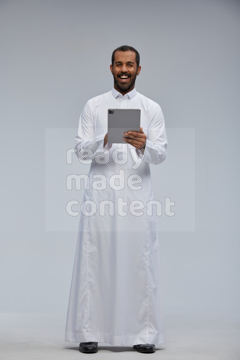 Saudi man wearing Thob standing working on tablet on Gray background